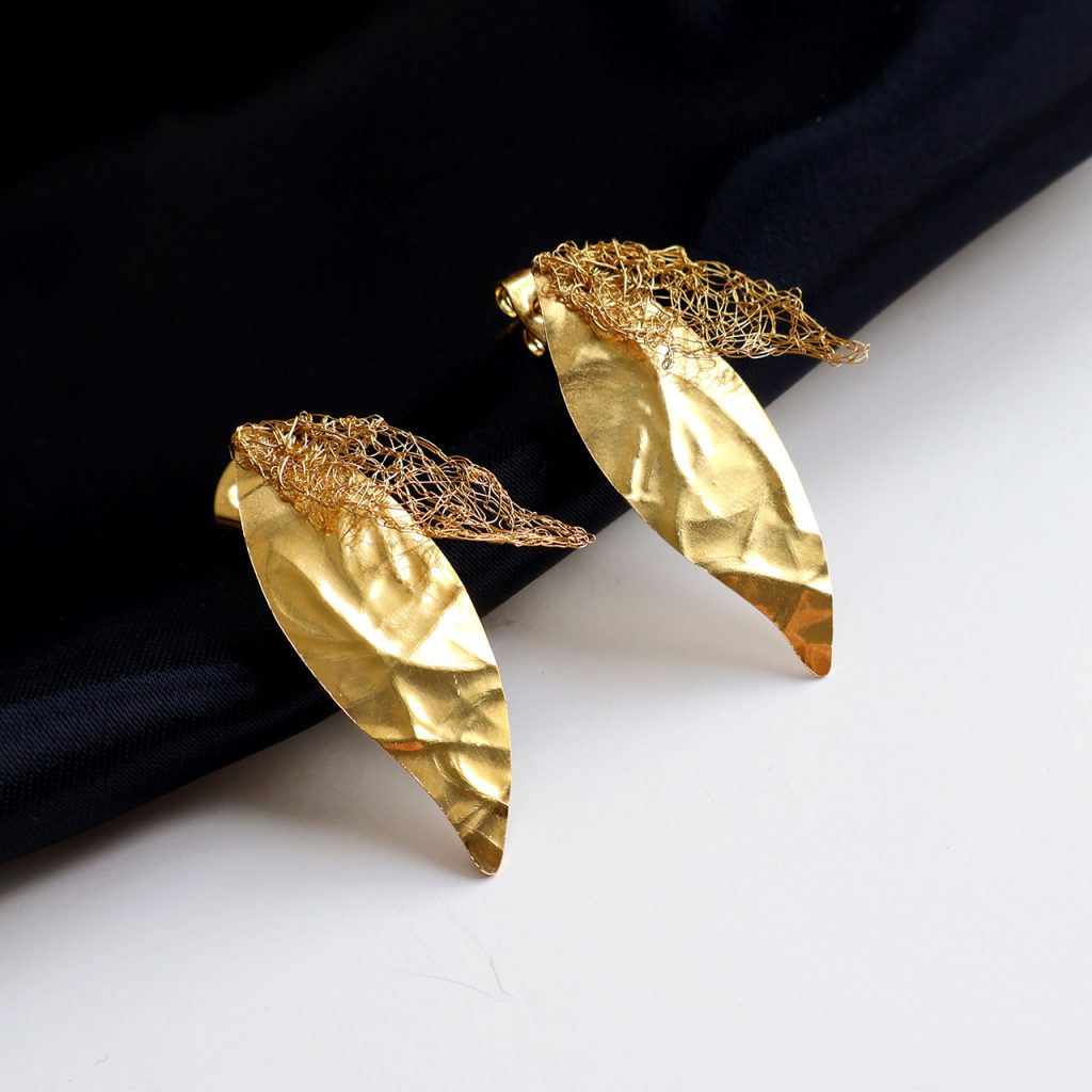 Les boucles d'oreilles Folÿs 1 sont des bijoux uniques brodés à la main par la marque Marie Archambaud. Elles reposent délicatement sur un tissu bleu marine, mettant en valeur l'éclat doré du métal. Elles sont composées de deux feuilles légères, une brodée au fil fin, et une en plaque de métal texturée, évoquant une nature poétique.