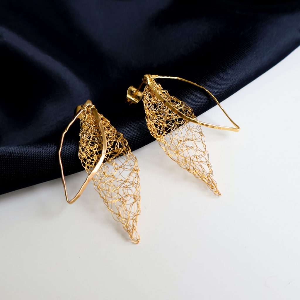 Les boucles d'oreilles Folÿs 4 sont des bijoux uniques brodés à la main par la marque Marie Archambaud. Elles reposent délicatement sur un tissu bleu marine, mettant en valeur l'éclat doré du métal. Elles sont composées de deux feuilles légères évoquant une nature délicate.