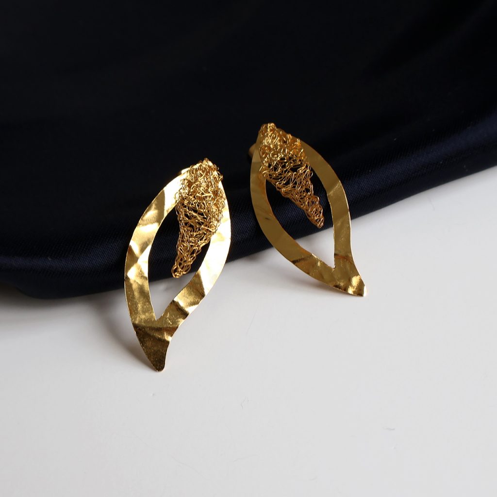 Les boucles d'oreilles Folÿs 5 sont des bijoux uniques brodés à la main par la marque Marie Archambaud. Elles reposent délicatement sur un tissu bleu marine, mettant en valeur l'éclat doré du métal. Sous forme de feuille légère, les boucles sont formées d'une plaque de métal texturée et de délicats fils brodés en leur centre.