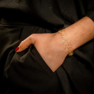 Poignet de femme sublimé par un bracelet en maille métallique unique confectionné à la main. La main est dans une poche de pantalon noir et le bijou fin est en coloris or jaune.