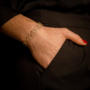 Main portant un bracelet unique conçu à la main par une technique de maille métallique. La main est dans une poche de pantalon noir et le bijou est en coloris or clair appelé "champagne".