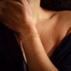Main portant le bracelet Lumio, couleur or jaune. C'est un bijou unique brodé à la main par une technique de maille métallique ajustable.