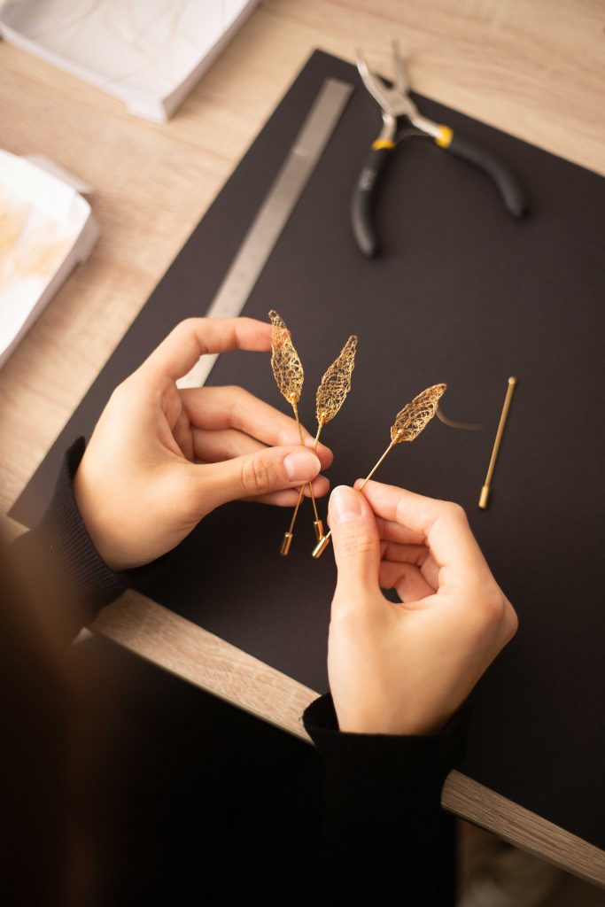 Les mains de la brodeuse m&eacute;talliques Marie Archambaud tiennent trois boutonni&egrave;res Aron, en cuivre dor&eacute; &agrave; l'or fin.