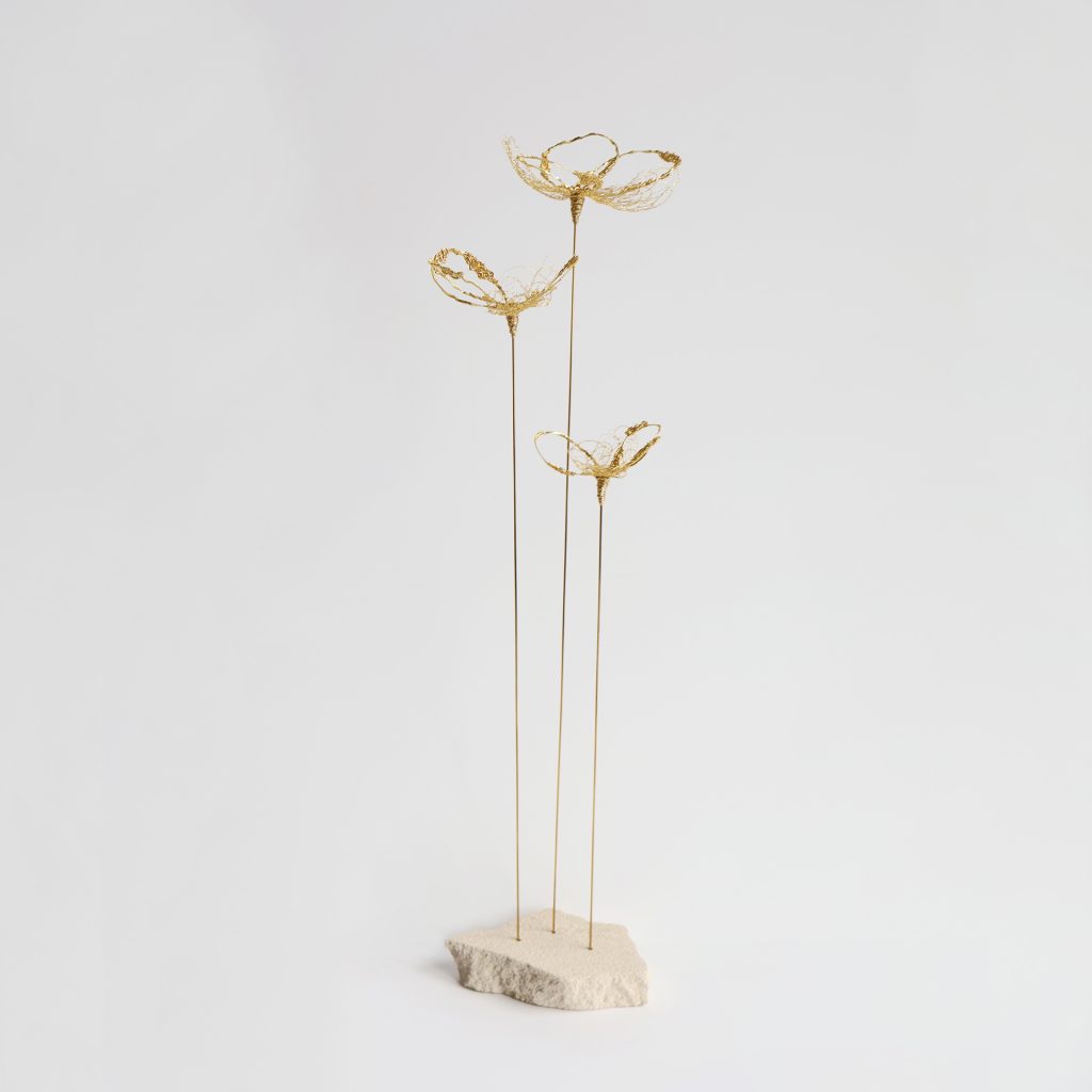 Sculpture Coccum sur fond gris. Trois tiges métalliques surmontées de fleurs brodées au fil d'or et piquées sur pierre de tufeau.