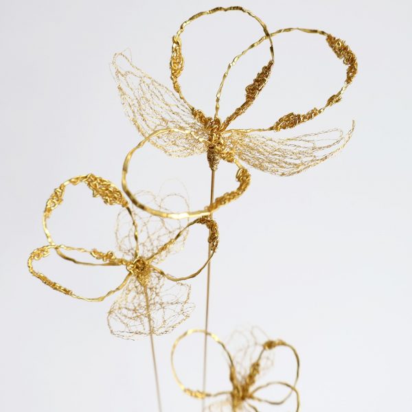 Sculpture Coccum sur fond gris. Trois tiges de laiton surmontées de fleurs brodées au fil d'or.