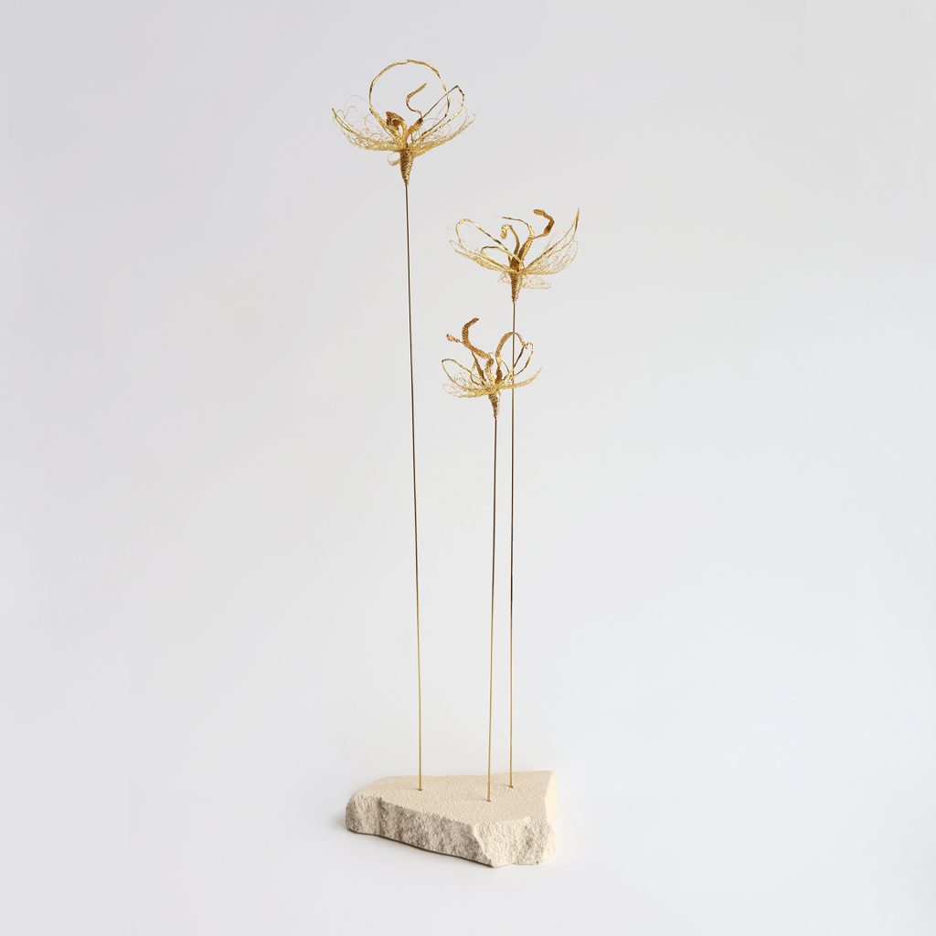 Sculpture Iridis sur fond gris. Trois tiges métalliques surmontées de fleurs brodées au fil d'or et piquées sur pierre de tufeau.