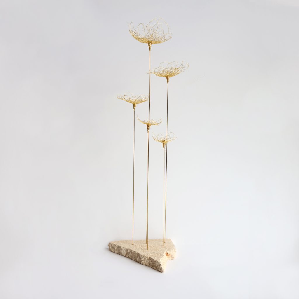 Sculpture Pavon sur fond gris. Cinq tiges métalliques surmontées de fleurs brodées au fil d'or et piquées sur pierre de tufeau.