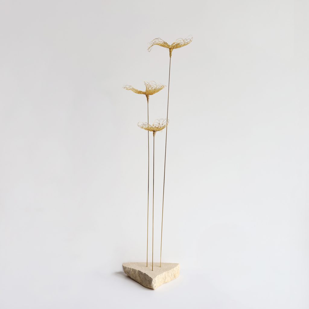 Sculpture Trifo sur fond gris. Trois tiges métalliques surmontées de fleurs brodées au fil d'or et piquées sur pierre de tufeau.