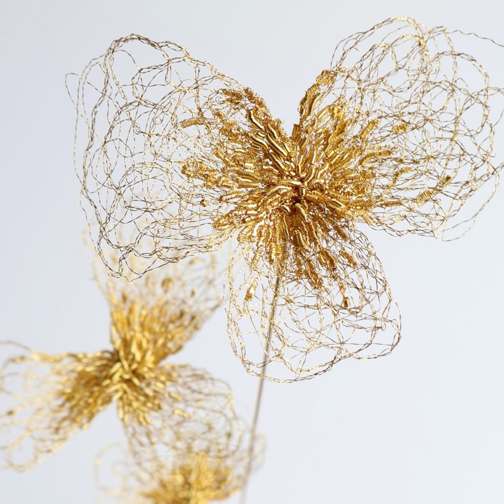 Détails de la sculpture Trifo sur fond gris. Trois tiges de laiton sont surmontées de fleurs de trèfle brodées au fil d'or.